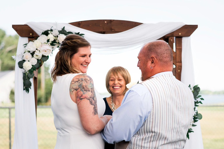 A bride with a tree tattoo on her arm smiling at her groom at the altar