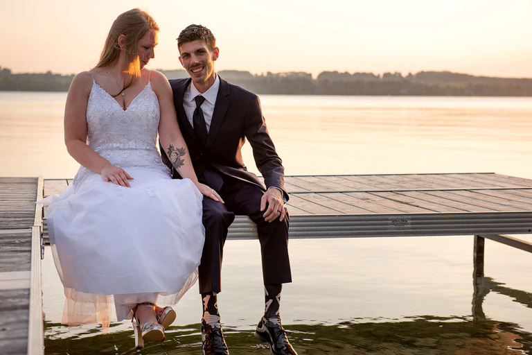 a married couple hold hands and sit on a dock on elk lake