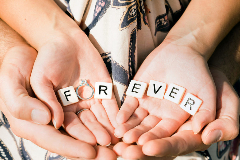 Couple's hands holding scrabble letters that spell Forever with the engagement ring as the O