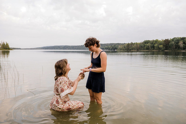one woman in a dress in the lake kneels as she proposes to her surprised partner