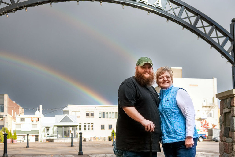 a mother and son stand in front of the arch with a double rainbow at city park