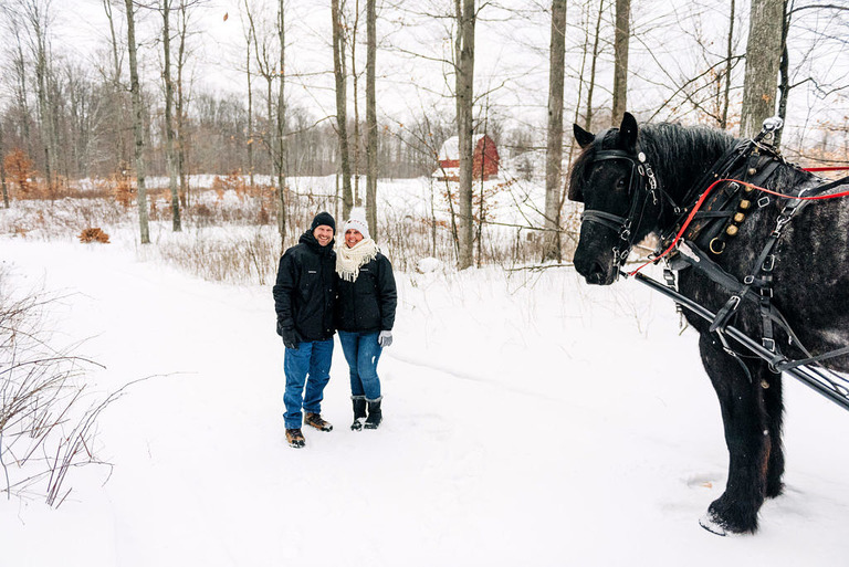 a couple stands in snow with a black horse at fantail farm
