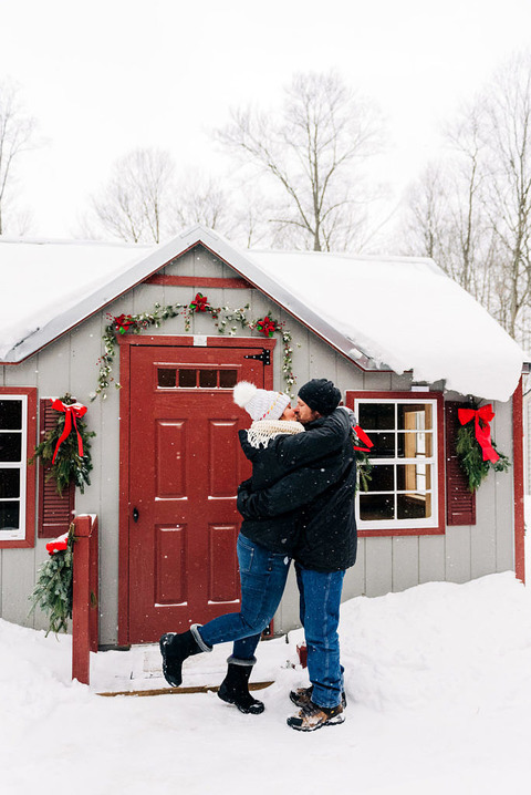 a newly engaged couple in snow kiss outside a decorated cabin at fantail farm