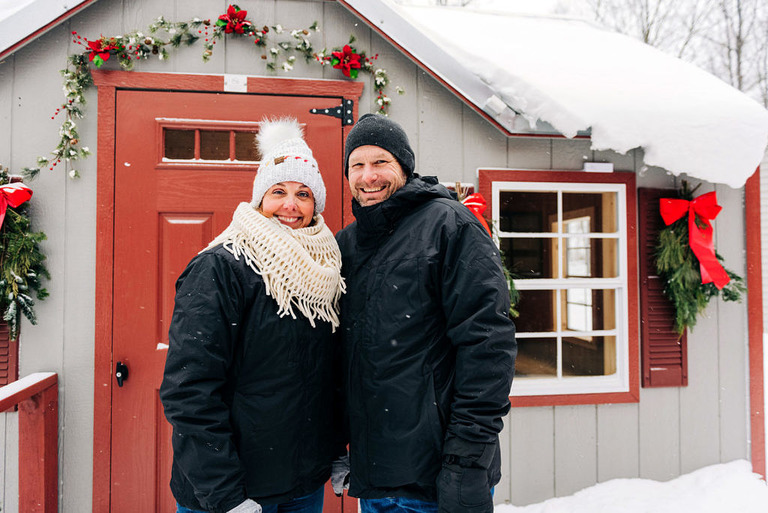 a newly engaged couple in snow stand outside a decorated cabin at fantail farm