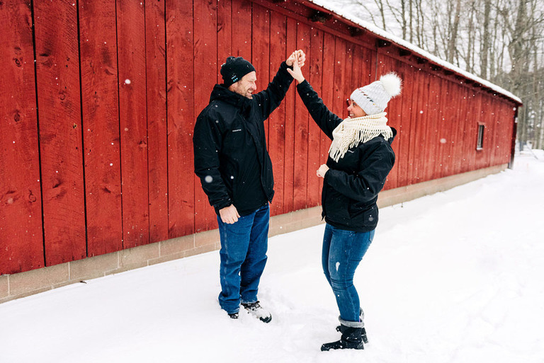 a newly engaged couple dance in the snow in front of a red barn