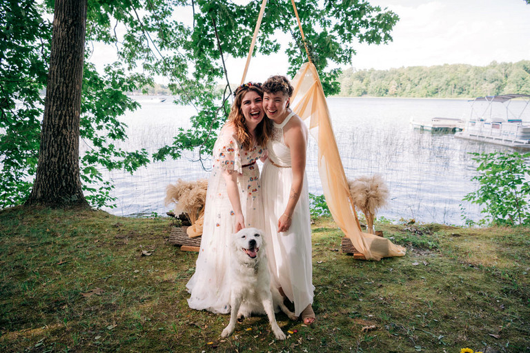 two brides and their dog smile in front of their wedding arch