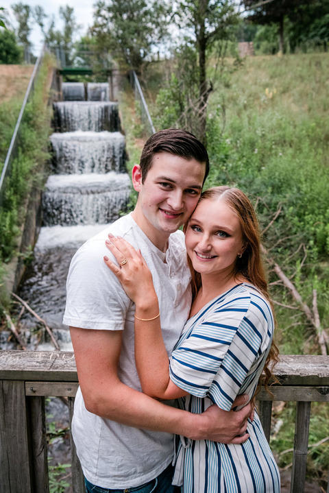 a man and woman embrace for engagement photos in front of the water steps at Mayfield Pond Park.