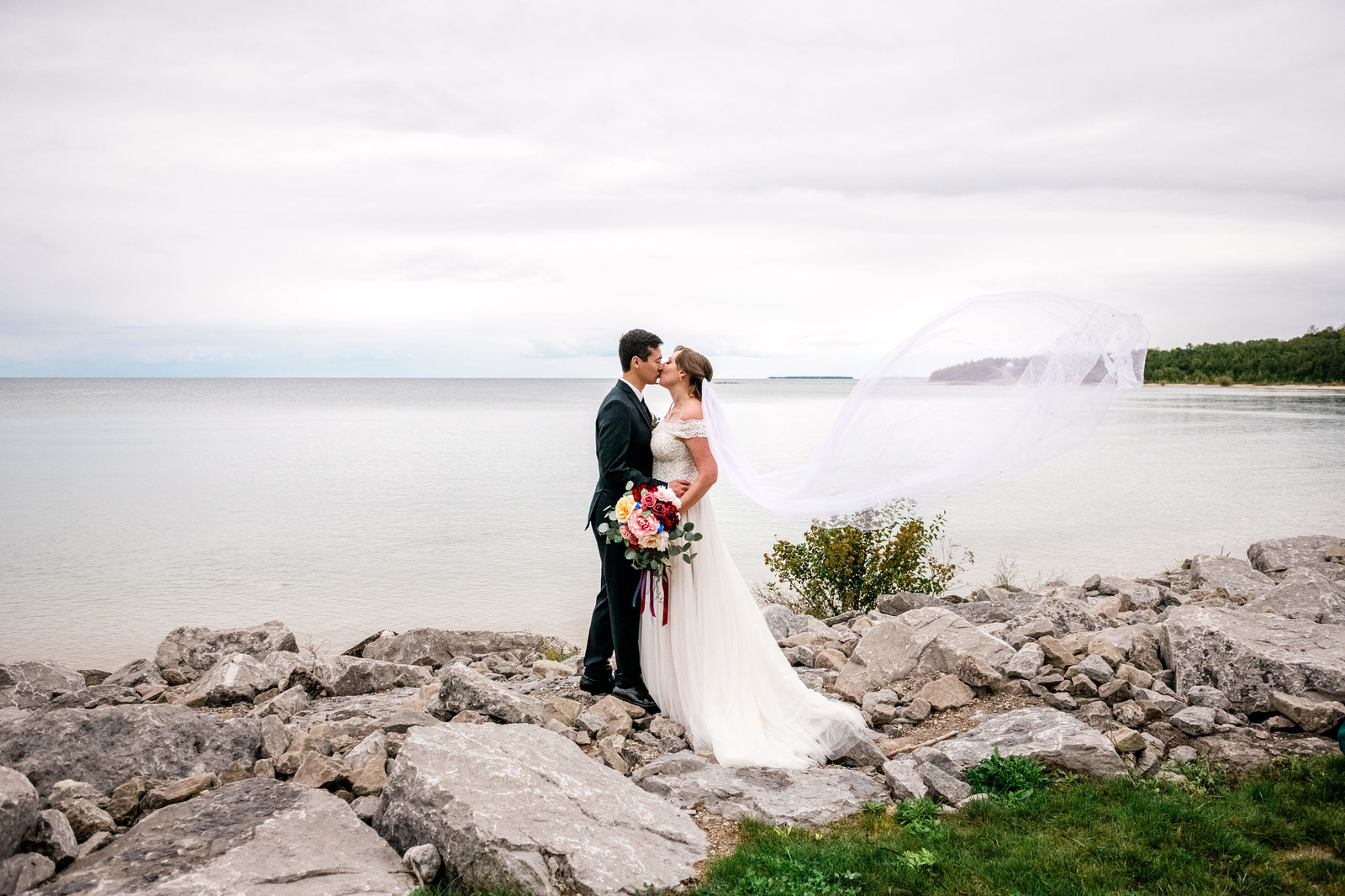 a couple in traditional wedding dress stand on rocks kissing by the lake with the veil flowing.