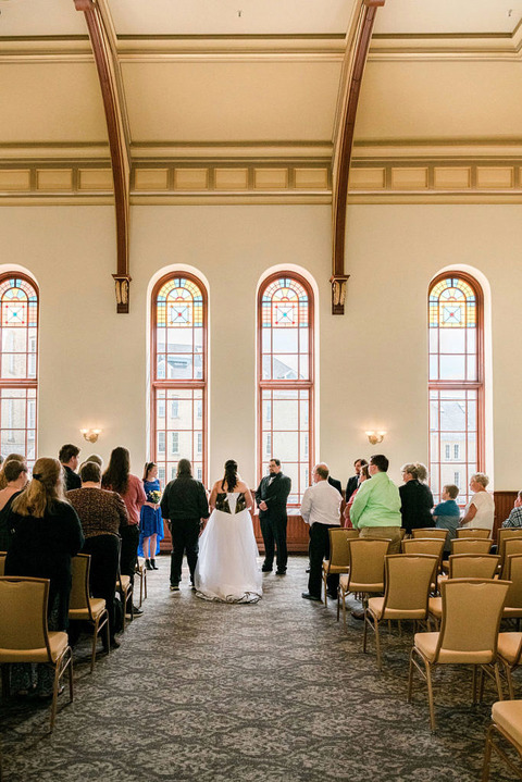 a bride and groom stand for their ceremony at Kirkbride Hall. Sun Streams in the ten foot stained glass windows.