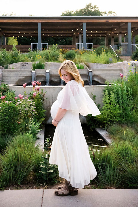 a woman in a white flutter-sleeve dress and cowboy boots stands in front of a fountain for pregnancy photos.