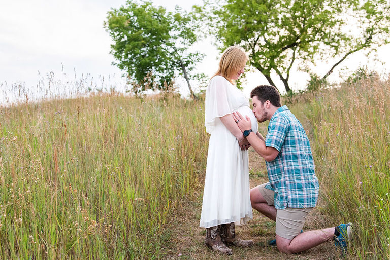 A man is kneeling and kissing a baby bump for pregnancy photos outdoors in a field in Traverse City.