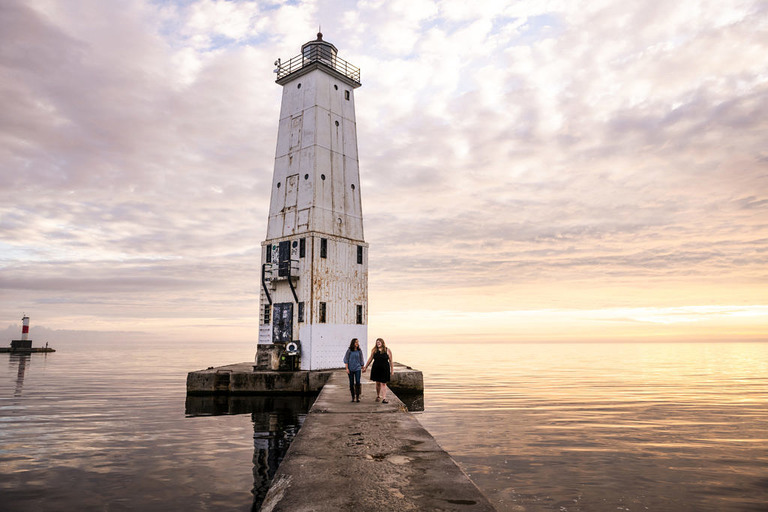 A couple walks hand in hand in front of a lighthouse in northern Michigan for photos.