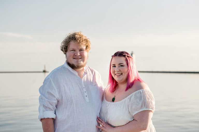 a couple who are engaged are smiling and facing the camera in front of the Frankfort lighthouse.