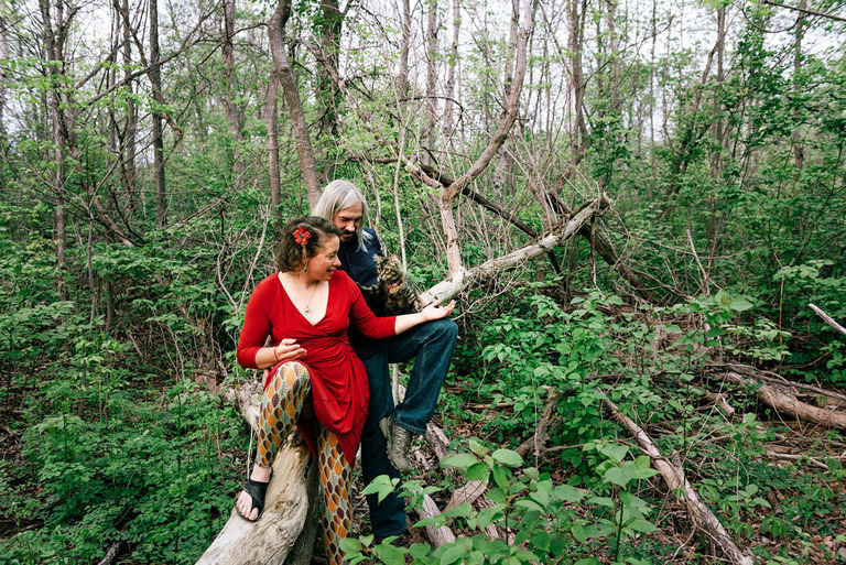 a cat, a woman in red, and a man all sit on a fallen tree branch for engagement photos