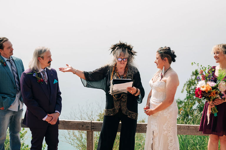 the officiant wearing black and gold with a feather crown invites the wedding couple to kiss on Sleeping Bear Dune.