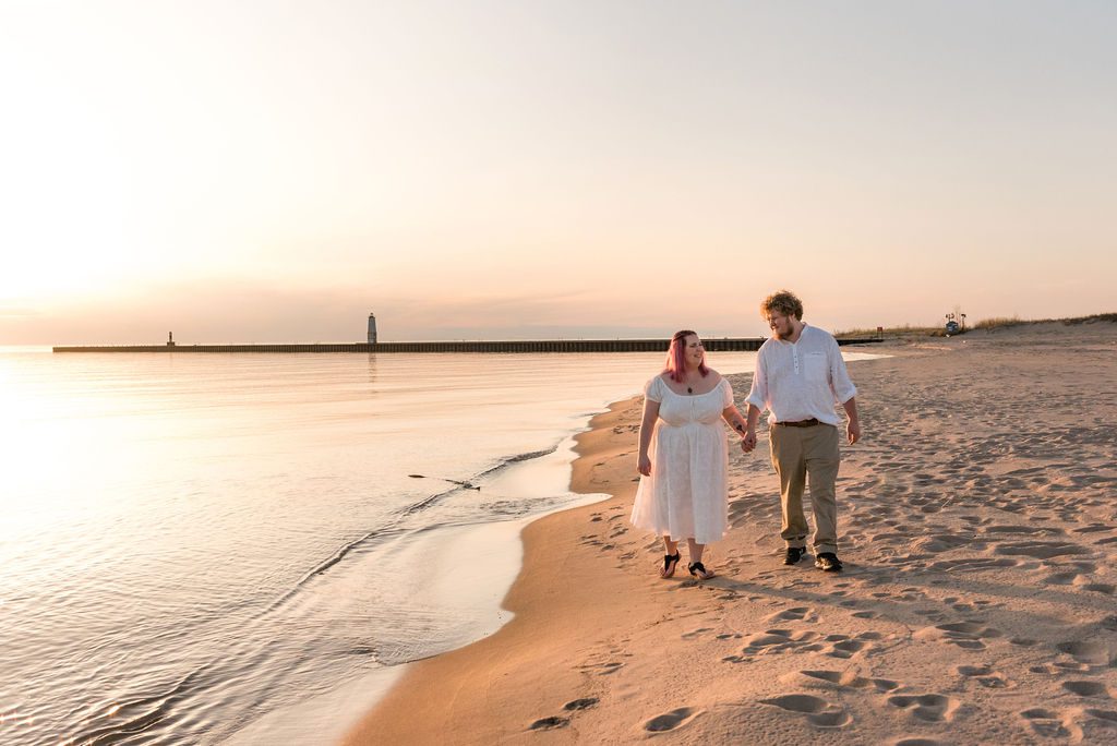 a couple in white walks hand in hand on Elberta beach at sunset.