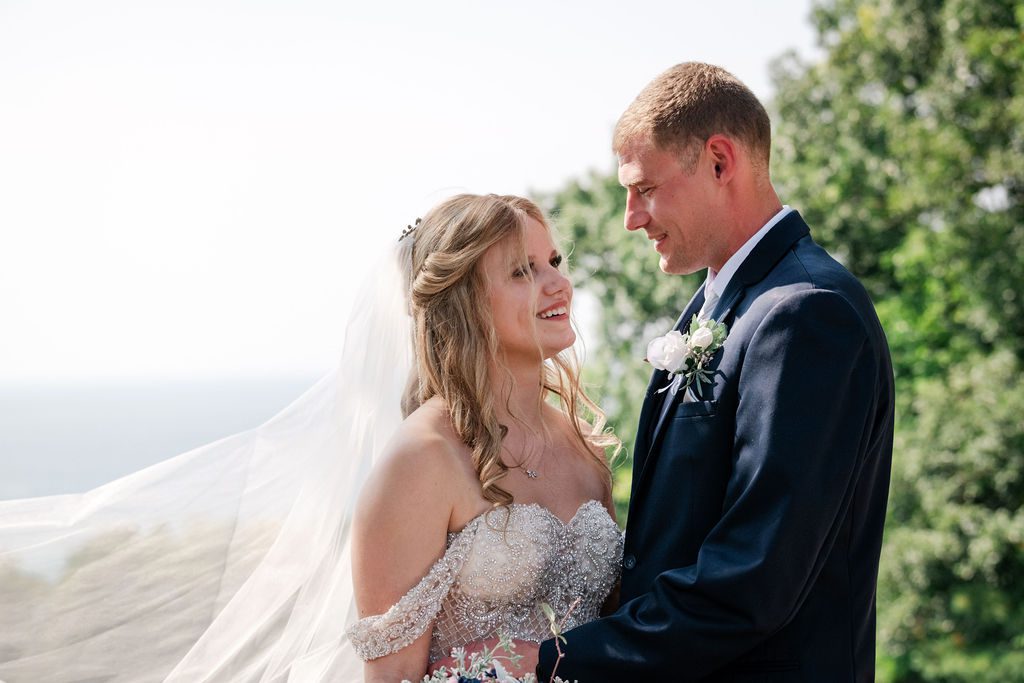 a bride wearing a beaded off the shoulder gown looking at her husband at the homestead.