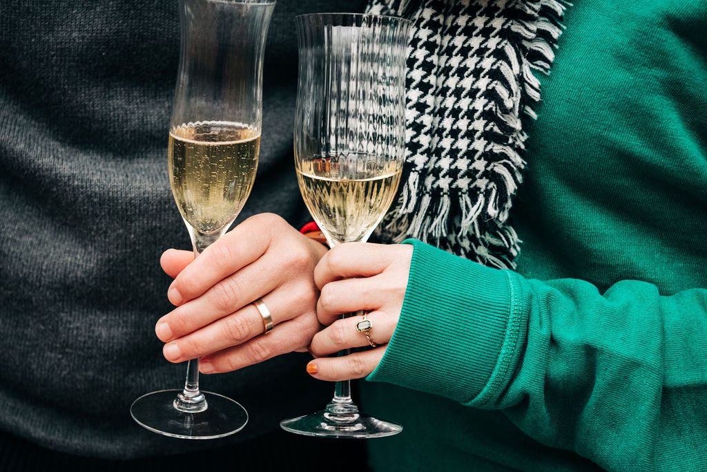 two hands of a surprise proposal couple showing their rings with glasses of champagne in the frame.