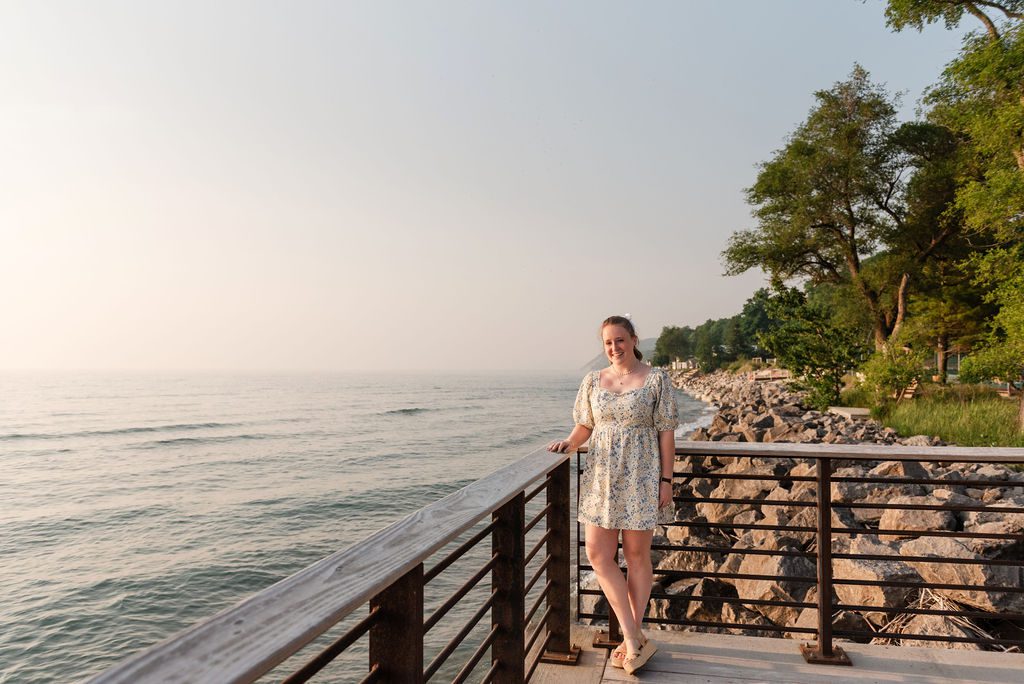 A young woman poses for senior portraits with wildfire haze in the sky along the shore of Lake Michigan at Camp Arcadia.