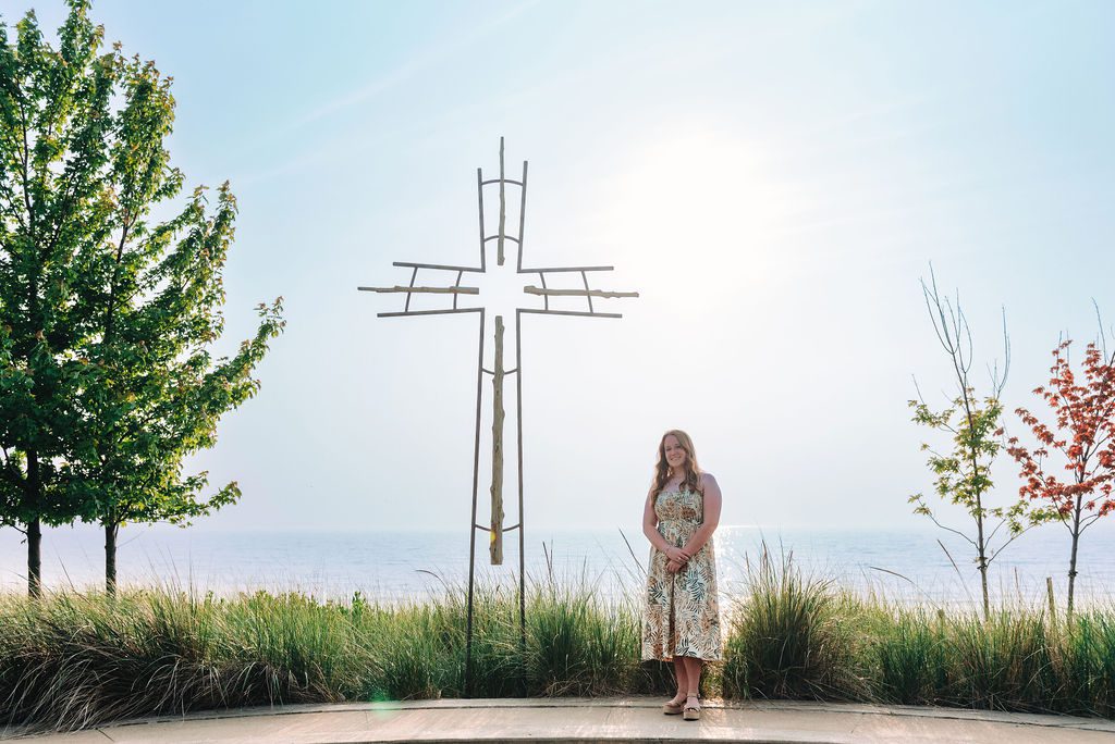 A young woman poses for senior portraits with wildfire haze in the sky along the shore of Lake Michigan at Camp Arcadia and the cross.